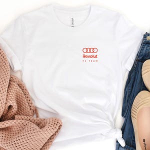 May include: A white t-shirt with a red Audi logo and the text "Revolut F1 TEAM". The shirt is styled with blue jeans, beige flats, and a pink knitted throw.