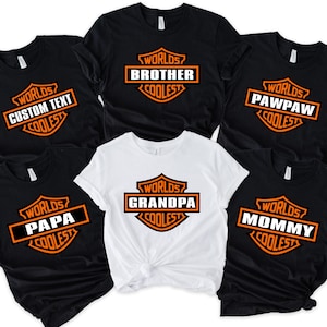 May include: A collection of black and white t-shirts with orange and white text. The shirts feature the phrase "Worlds Coolest" above various family titles like "Brother", "Grandpa", and "Mommy". The shirts are short-sleeved.