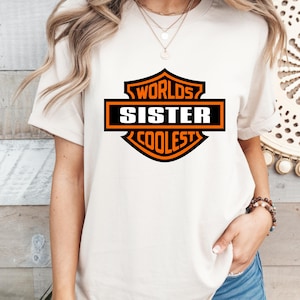 May include: Cream-colored t-shirt featuring a black and orange graphic that reads "Worlds Sister Coolest". The design is in a shield shape. The shirt has short sleeves and a crew neck.