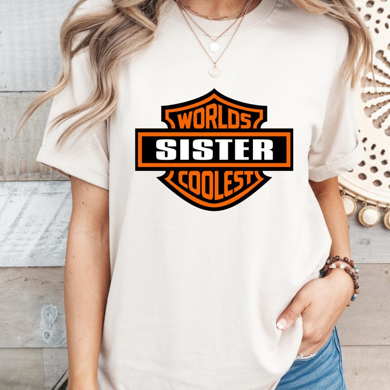 May include: Cream-colored t-shirt featuring a black and orange graphic that reads "Worlds Sister Coolest". The design is in a shield shape. The shirt has short sleeves and a crew neck.