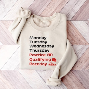 May include: A cream-colored sweatshirt with the days of the week printed in black and red. The text includes "Monday" through "Thursday", "Practice", "Qualifying", and "Raceday". A coffee cup and race car graphic are included.
