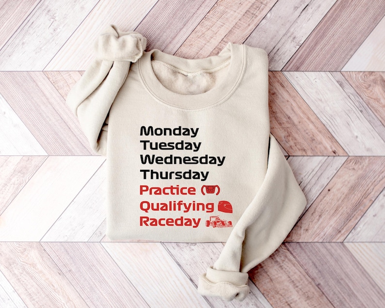 May include: A cream-colored sweatshirt with the days of the week printed in black and red. The text includes "Monday" through "Thursday", "Practice", "Qualifying", and "Raceday". A coffee cup and race car graphic are included.