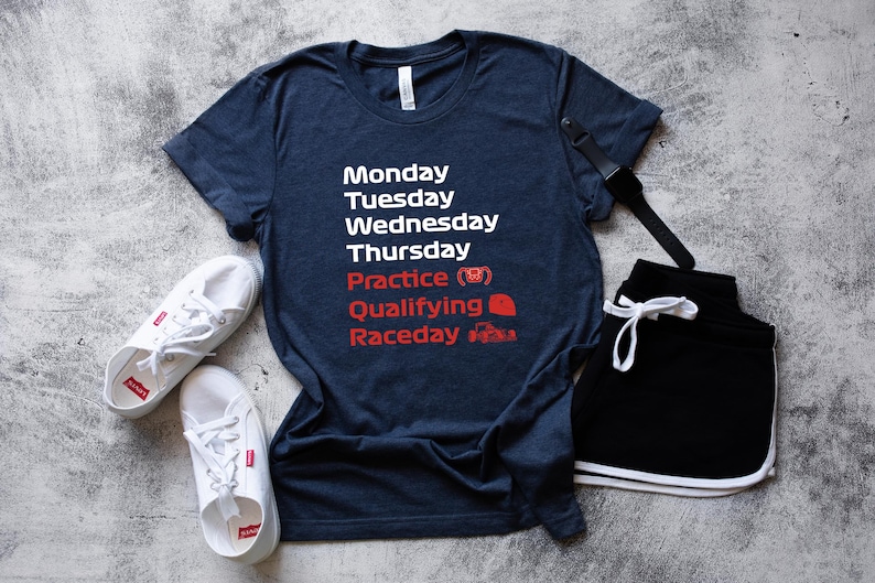 May include: A navy blue t-shirt with white text listing days of the week and racing terms in red. The shirt is paired with black shorts, white sneakers, and a black watch. The text includes "Practice", "Qualifying", and "Raceday".