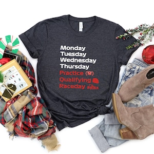 May include: Dark gray t-shirt with white and red text. The text lists days of the week, practice, qualifying, and raceday. The shirt is paired with blue jeans and tan boots. A gift box, scarf, and Christmas ornaments are also in the image.