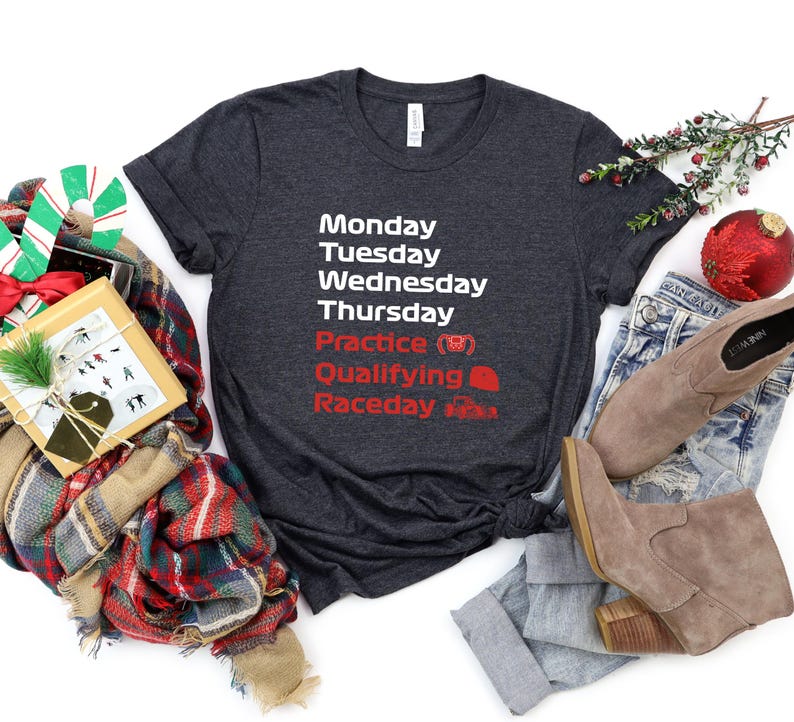 May include: Dark gray t-shirt with white and red text. The text lists days of the week, practice, qualifying, and raceday. The shirt is paired with blue jeans and tan boots. A gift box, scarf, and Christmas ornaments are also in the image.