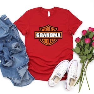 May include: A red t-shirt with the text "Worlds Grandma Coolest" in a black and orange design. The image also includes a denim shirt, white sneakers, and a bouquet of pink roses.