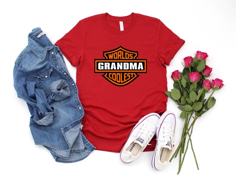 May include: A red t-shirt with the text "Worlds Grandma Coolest" in a black and orange design. The image also includes a denim shirt, white sneakers, and a bouquet of pink roses.