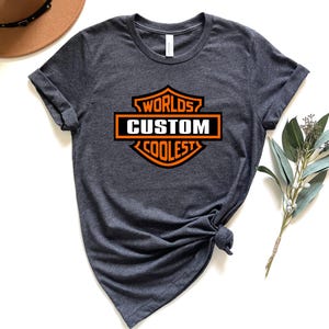 May include: Dark gray t-shirt with a crew neck and rolled sleeves. The shirt features an orange and white graphic that reads "Worlds Custom Coolest" in a shield design. A brown hat and greenery are in the background.