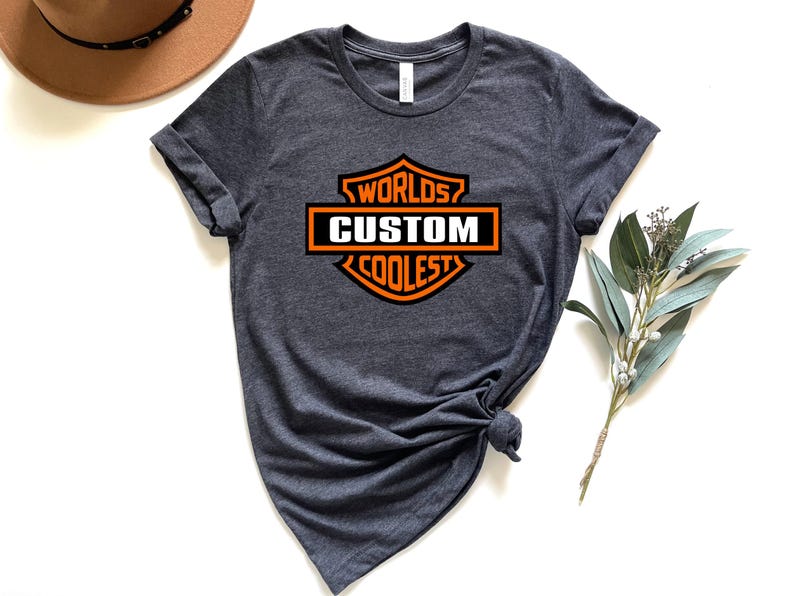 May include: Dark gray t-shirt with a crew neck and rolled sleeves. The shirt features an orange and white graphic that reads "Worlds Custom Coolest" in a shield design. A brown hat and greenery are in the background.