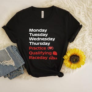 May include: Black t-shirt with white and red text listing days of the week and racing-related terms. Includes a pair of folded blue jeans and a yellow sunflower. The text includes "Practice", "Qualifying", and "Raceday".