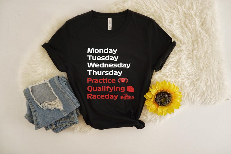 May include: Black t-shirt with white and red text listing days of the week and racing-related terms. Includes a pair of folded blue jeans and a yellow sunflower. The text includes "Practice", "Qualifying", and "Raceday".