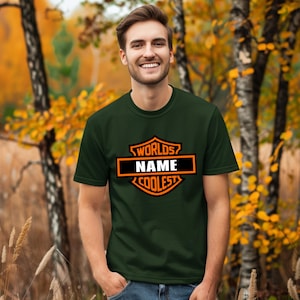 May include: Dark green t-shirt with a black and orange graphic that reads "WORLDS NAME COOLEST". The t-shirt is short-sleeved and is being worn by a person. The background is a blurred outdoor scene with trees and fall foliage.
