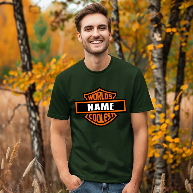 May include: Dark green t-shirt with a black and orange graphic that reads "WORLDS NAME COOLEST". The t-shirt is short-sleeved and is being worn by a person. The background is a blurred outdoor scene with trees and fall foliage.