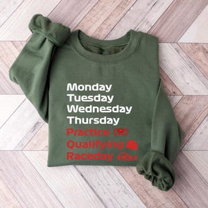 May include: An olive green sweatshirt with white text listing the days of the week, and red text for "Practice", "Qualifying", and "Raceday". The sweatshirt is folded on a light brown wooden surface.