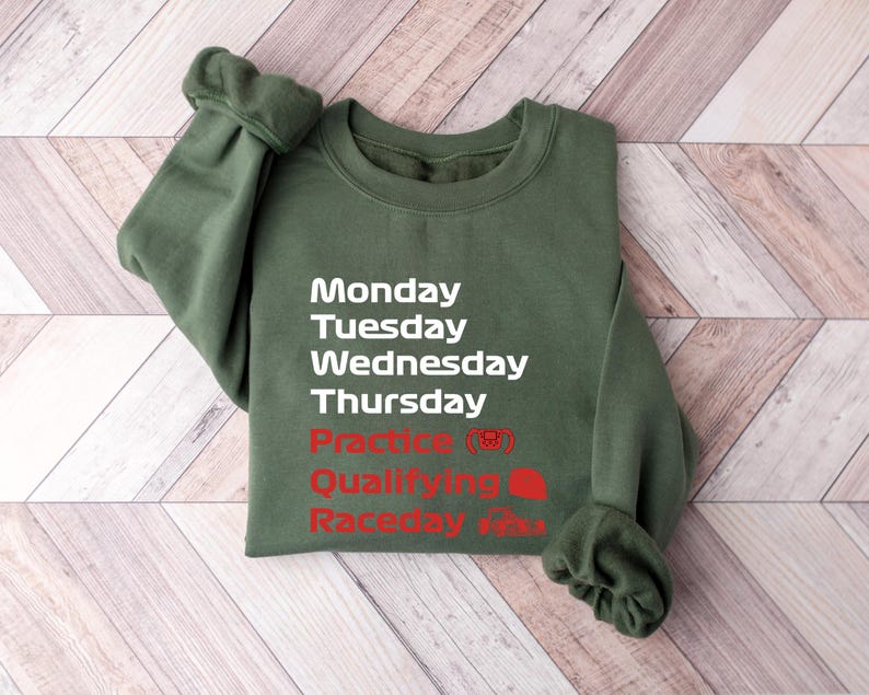 May include: An olive green sweatshirt with white text listing the days of the week, and red text for "Practice", "Qualifying", and "Raceday". The sweatshirt is folded on a light brown wooden surface.