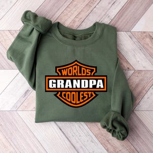 May include: An olive green sweatshirt with the words "Worlds Grandpa Coolest" in a black, orange, and white graphic. The sweatshirt is folded on a light brown and white wooden surface.