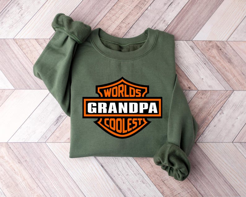 May include: An olive green sweatshirt with the words "Worlds Grandpa Coolest" in a black, orange, and white graphic. The sweatshirt is folded on a light brown and white wooden surface.