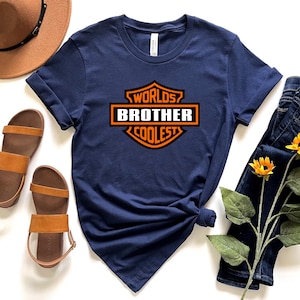 May include: Navy blue t-shirt with the text "Worlds Coolest Brother" in an orange and white design. The shirt is styled with brown sandals, a brown hat, and blue jeans with sunflowers.