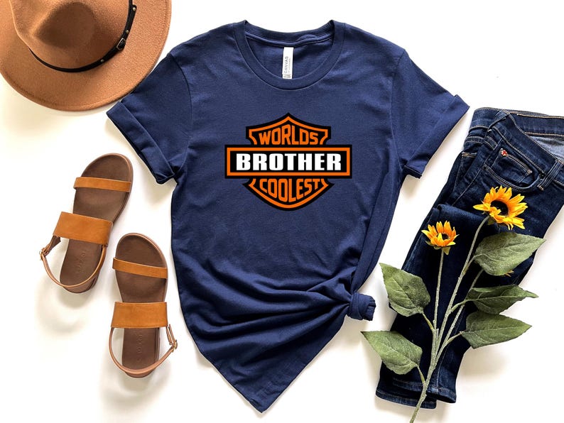 May include: Navy blue t-shirt with the text "Worlds Coolest Brother" in an orange and white design. The shirt is styled with brown sandals, a brown hat, and blue jeans with sunflowers.