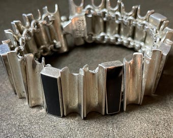 May include: A silver-toned bracelet with a rectangular design and two black rectangular insets. The bracelet is made of connected rectangular links with a textured, ridged design. The clasp is visible and the bracelet is on a gray surface.