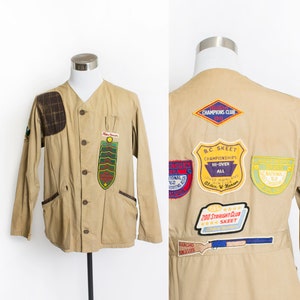 May include: A tan canvas jacket with a plaid patch on the left shoulder and several embroidered patches on the back. The patches feature the text "Champions Club", "B.C. Skeet Championships", "Hi-Over All", "200 Straight Club Skeet", and "Rancho Angelis".