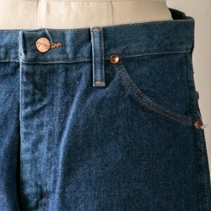Puede incluir: Primer plano de la cintura y el bolsillo delantero de un par de pantalones vaqueros de color azul oscuro. Un bot&oacute;n de color cobre con la palabra "Wrangler" es visible en la cintura.