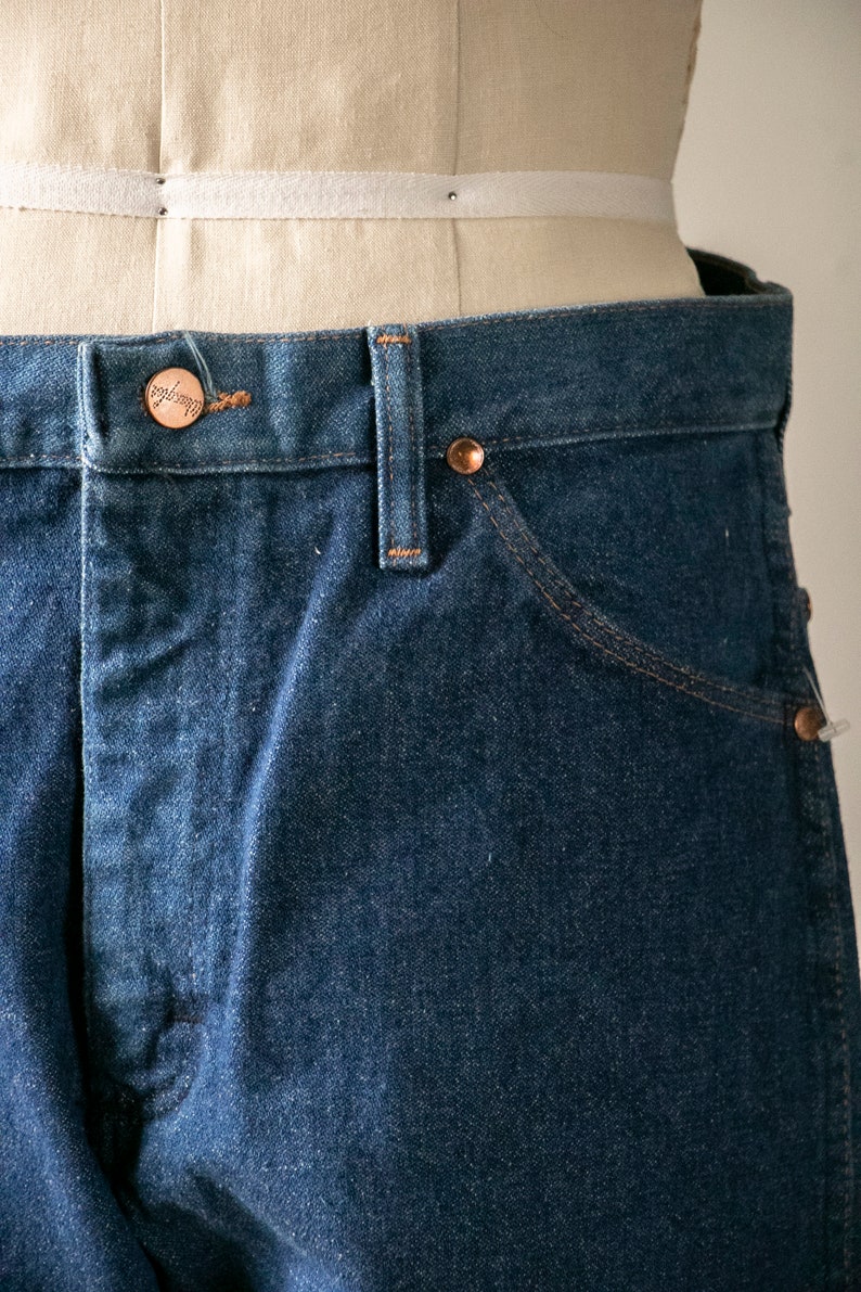 Puede incluir: Primer plano de la cintura y el bolsillo delantero de un par de pantalones vaqueros de color azul oscuro. Un bot&oacute;n de color cobre con la palabra "Wrangler" es visible en la cintura.