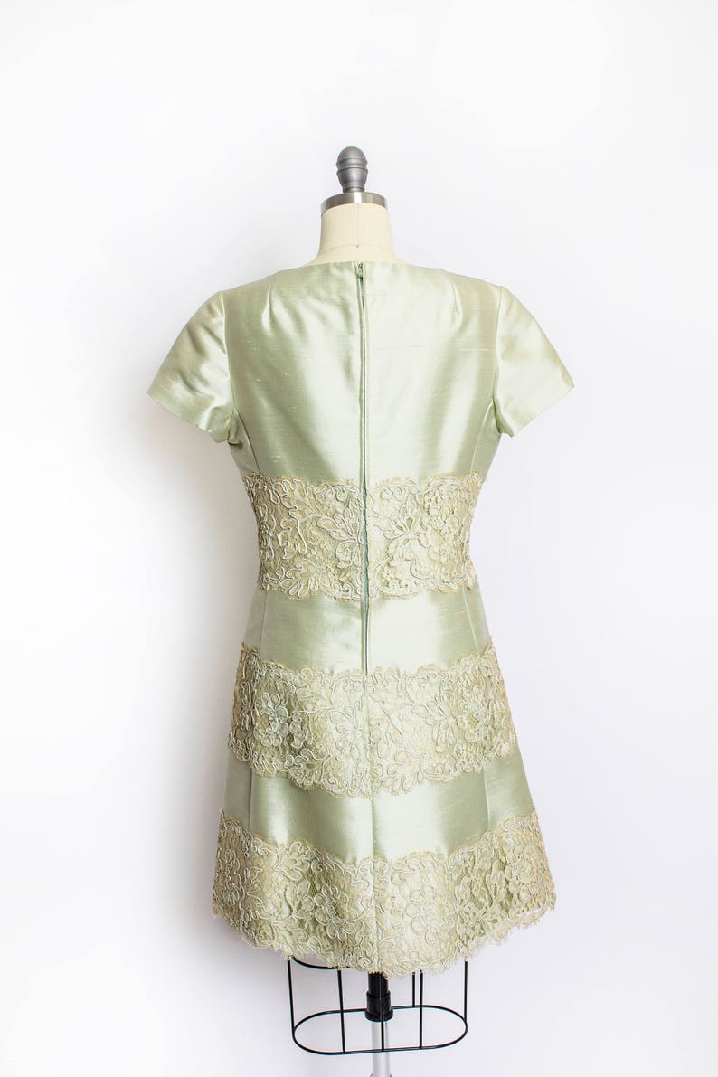 May include: A light green dress with a fitted bodice and a flared skirt. The dress is made of a silky fabric and features delicate lace trim around the neckline, waist, and hem.