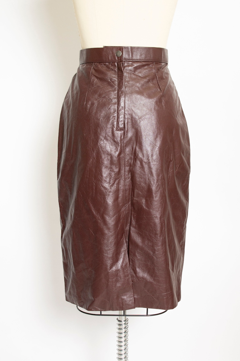 May include: A brown leather pencil skirt displayed on a mannequin. The skirt has a high waist with a button closure and a back slit. The leather has a slightly wrinkled texture.