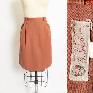 May include: A brown wool skirt with a high waist and a straight silhouette. The skirt is made in Italy and is labeled "Gucci" with the text "100% LANA / WOOL / LAINE" and "Made in Italy".