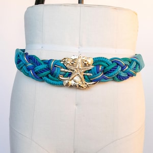 Pu&ograve; includere: Una cintura intrecciata nei toni del verde acqua e blu, con dettagli dorati e una fibbia a forma di stella marina dorata. La cintura &egrave; esposta su un manichino, evidenziando il suo tema nautico e il suo design.