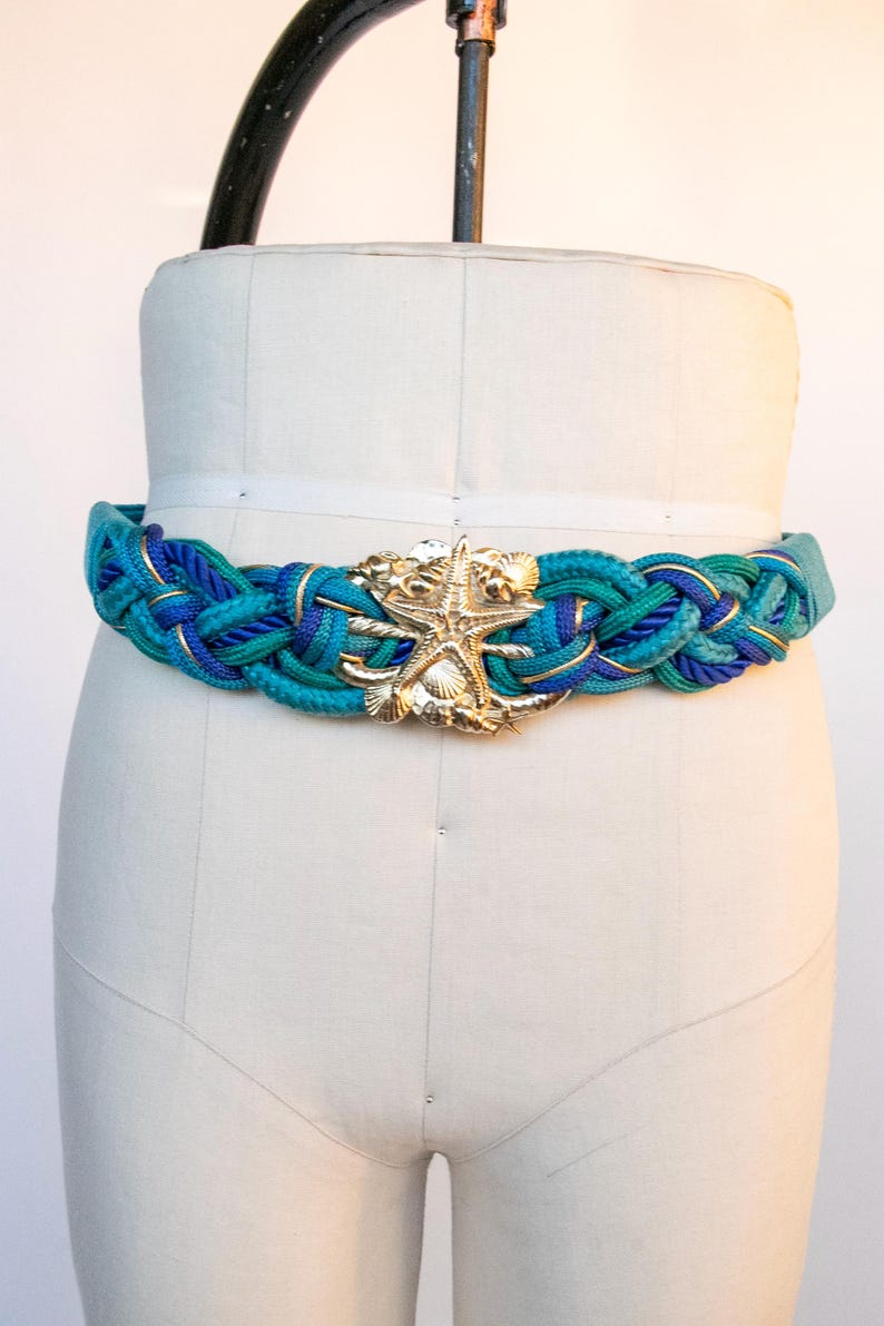 Pu&ograve; includere: Una cintura intrecciata nei toni del verde acqua e blu, con dettagli dorati e una fibbia a forma di stella marina dorata. La cintura &egrave; esposta su un manichino, evidenziando il suo tema nautico e il suo design.