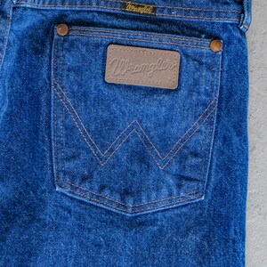 May include: A pair of blue denim jeans with a back pocket featuring a brown leather patch with the word "Wrangler" in white lettering.