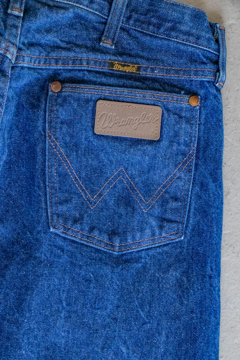 May include: A pair of blue denim jeans with a back pocket featuring a brown leather patch with the word "Wrangler" in white lettering.