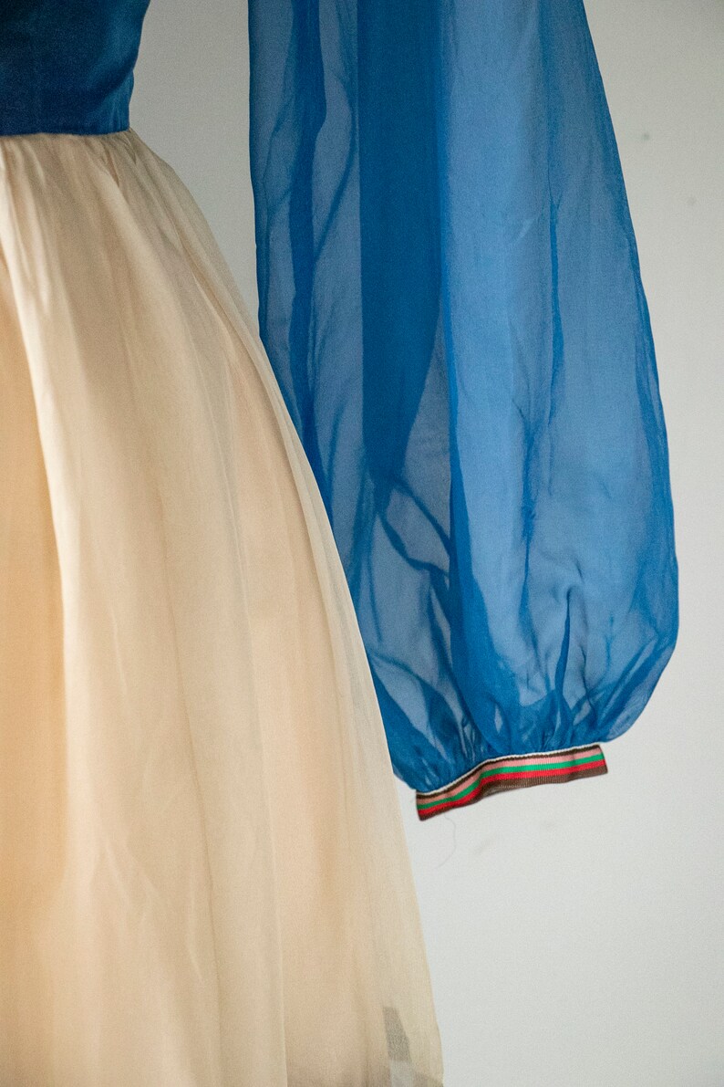 May include: A white tulle skirt with a blue sheer overlay. The overlay has a colorful ribbon trim.