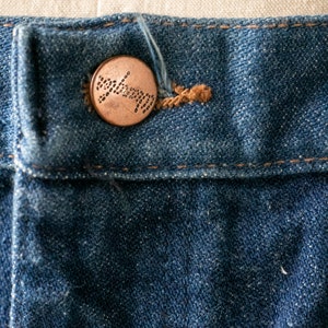 Puede incluir: Primer plano de un jean azul de mezclilla con un bot&oacute;n de cobre. El bot&oacute;n tiene un dise&ntilde;o circular con un patr&oacute;n de peque&ntilde;os puntos.