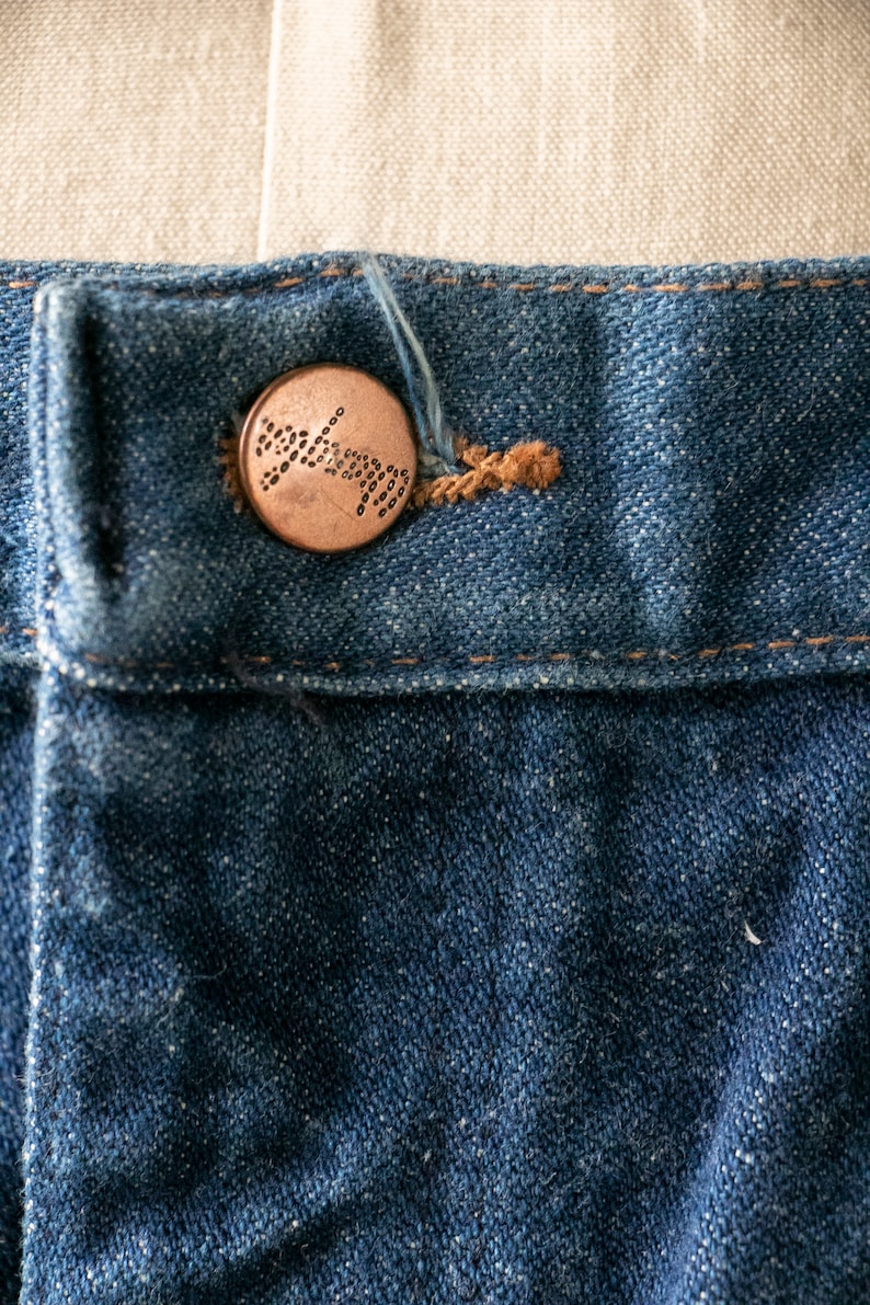 Puede incluir: Primer plano de un jean azul de mezclilla con un bot&oacute;n de cobre. El bot&oacute;n tiene un dise&ntilde;o circular con un patr&oacute;n de peque&ntilde;os puntos.