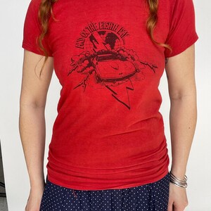 May include: Red t-shirt with a black graphic featuring a car, lightning bolts, and the text "And on the Eighth Day". The shirt is short-sleeved and has a crew neck. The model is wearing a navy blue polka dot skirt.