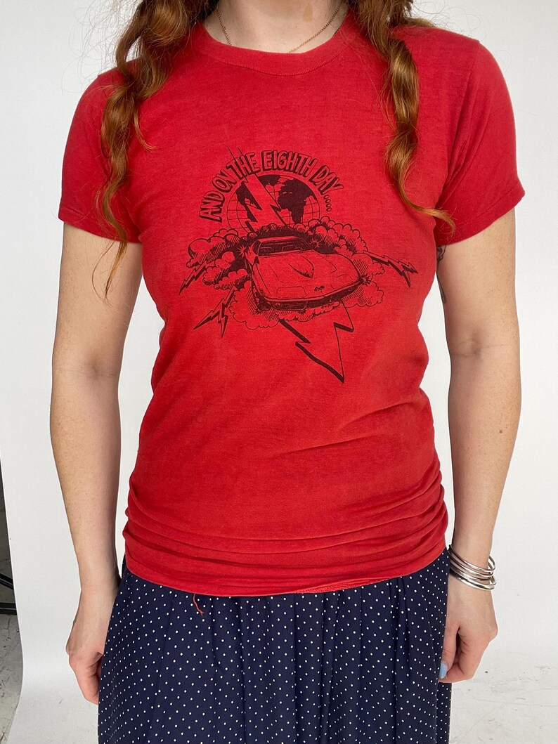 May include: Red t-shirt with a black graphic featuring a car, lightning bolts, and the text "And on the Eighth Day". The shirt is short-sleeved and has a crew neck. The model is wearing a navy blue polka dot skirt.