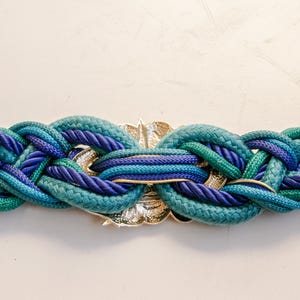 Pu&ograve; includere: Cintura intrecciata nei toni del verde acqua e blu con una chiusura in metallo color oro. La cintura &egrave; realizzata in spessa corda intrecciata, creando un accessorio strutturato e accattivante. Un'aggiunta elegante a qualsiasi guardaroba.