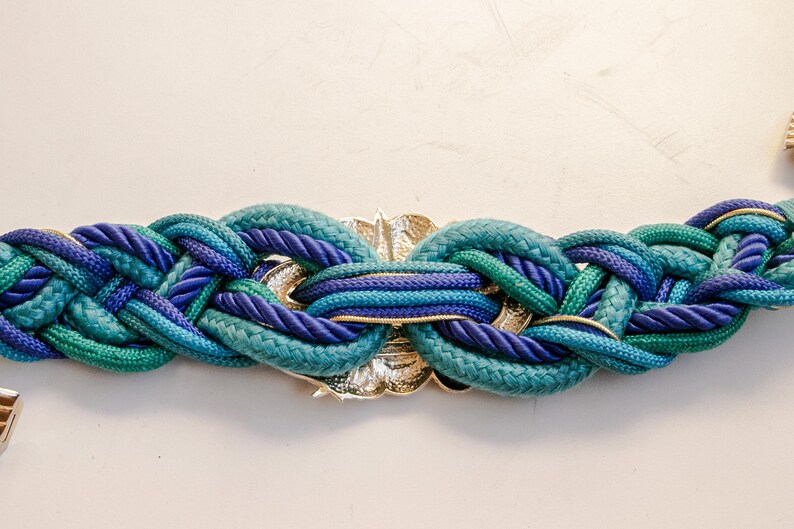 Pu&ograve; includere: Cintura intrecciata nei toni del verde acqua e blu con una chiusura in metallo color oro. La cintura &egrave; realizzata in spessa corda intrecciata, creando un accessorio strutturato e accattivante. Un'aggiunta elegante a qualsiasi guardaroba.