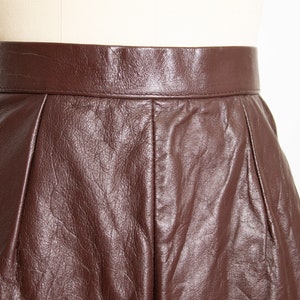 May include: Close-up of a brown leather skirt. The skirt has a high waist and a pleated design. The leather has a textured appearance and a rich, dark brown color. The skirt is displayed on a mannequin.