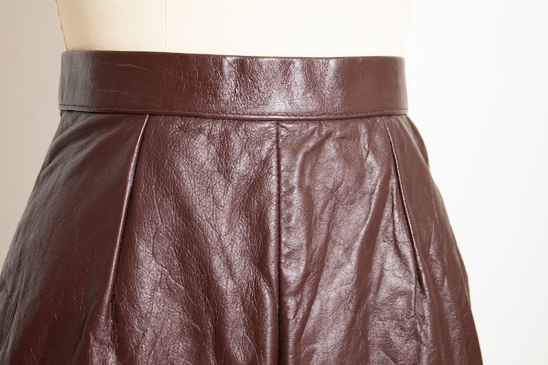 May include: Close-up of a brown leather skirt. The skirt has a high waist and a pleated design. The leather has a textured appearance and a rich, dark brown color. The skirt is displayed on a mannequin.