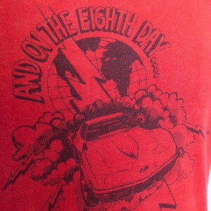 May include: Red t-shirt with a black graphic design. The design features a car, lightning bolt, and a globe with the text "AND ON THE EIGHTH DAY". The shirt is made of a soft material.