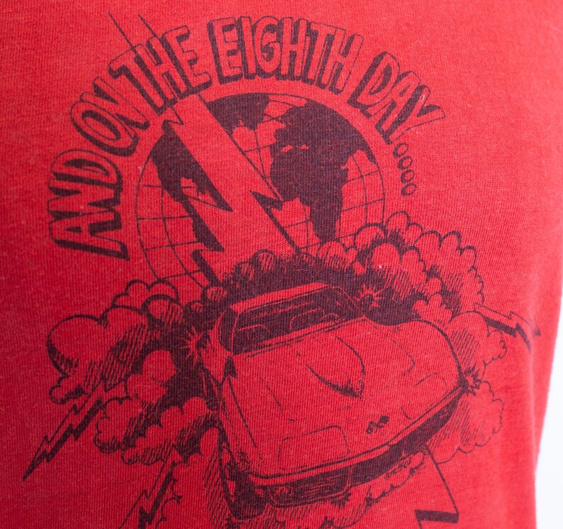 May include: Red t-shirt with a black graphic design. The design features a car, lightning bolt, and a globe with the text "AND ON THE EIGHTH DAY". The shirt is made of a soft material.