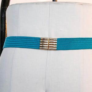 Pu&ograve; includere: Una cintura elastica turchese con una chiusura rettangolare color oro. La cintura &egrave; esposta su un manichino bianco. La chiusura &egrave; composta da due pezzi rettangolari che si collegano al centro. Accessorio moda.