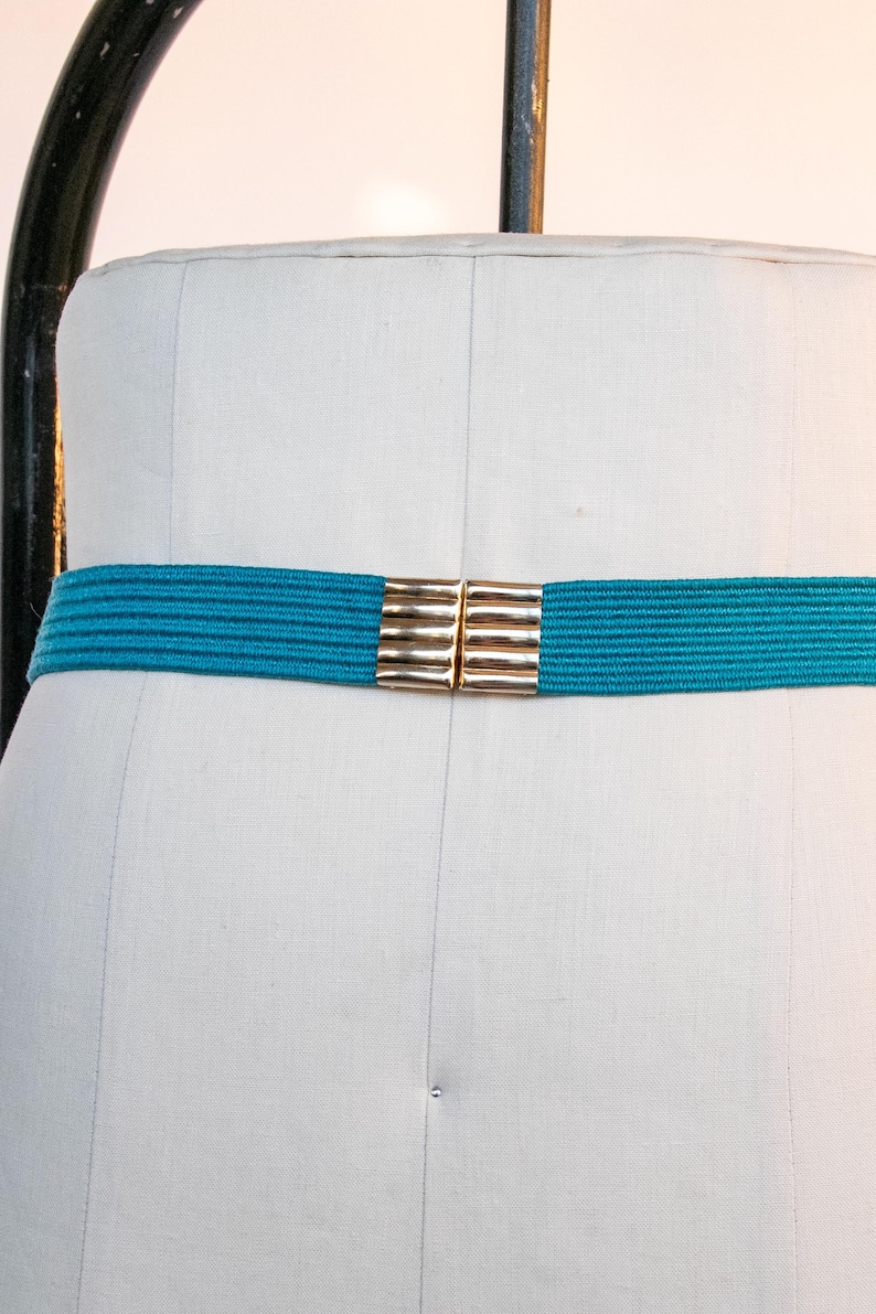 Pu&ograve; includere: Una cintura elastica turchese con una chiusura rettangolare color oro. La cintura &egrave; esposta su un manichino bianco. La chiusura &egrave; composta da due pezzi rettangolari che si collegano al centro. Accessorio moda.