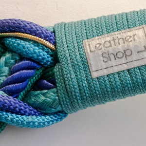 Pu&ograve; includere: Primo piano di una cintura intrecciata nelle tonalit&agrave; del blu e del verde acqua. La cintura presenta un anello color oro e un'etichetta con la scritta "Leather Shop". La sezione intrecciata &egrave; realizzata in materiale spesso e tessuto.