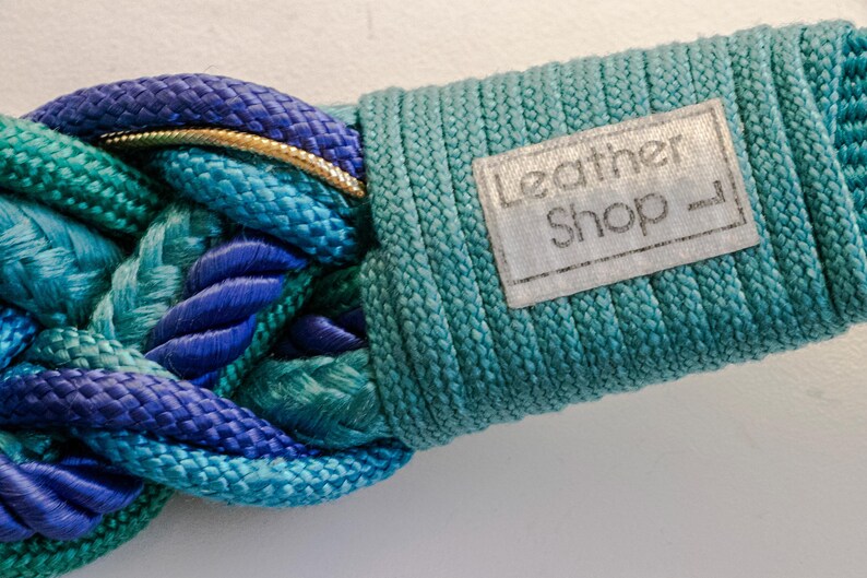 Pu&ograve; includere: Primo piano di una cintura intrecciata nelle tonalit&agrave; del blu e del verde acqua. La cintura presenta un anello color oro e un'etichetta con la scritta "Leather Shop". La sezione intrecciata &egrave; realizzata in materiale spesso e tessuto.