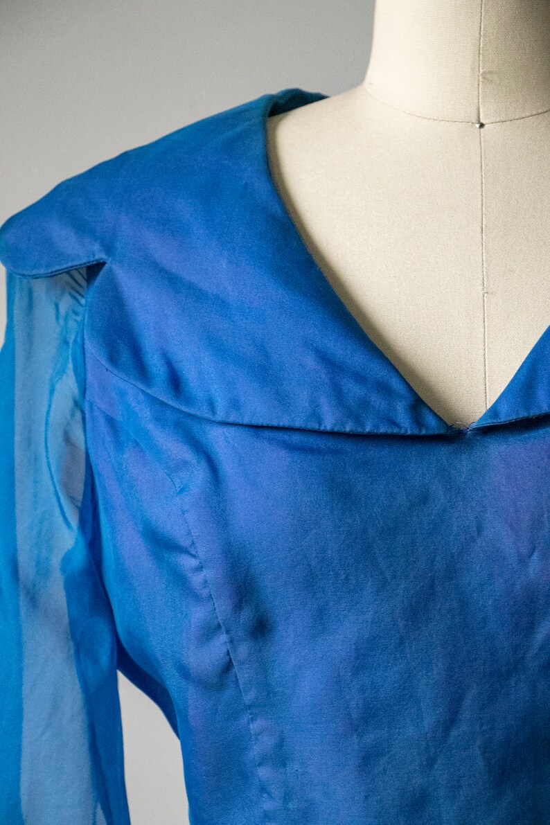 May include: A blue sheer blouse with a V-neck and long sleeves. The blouse has a collar that folds down and a gathered detail at the neckline.